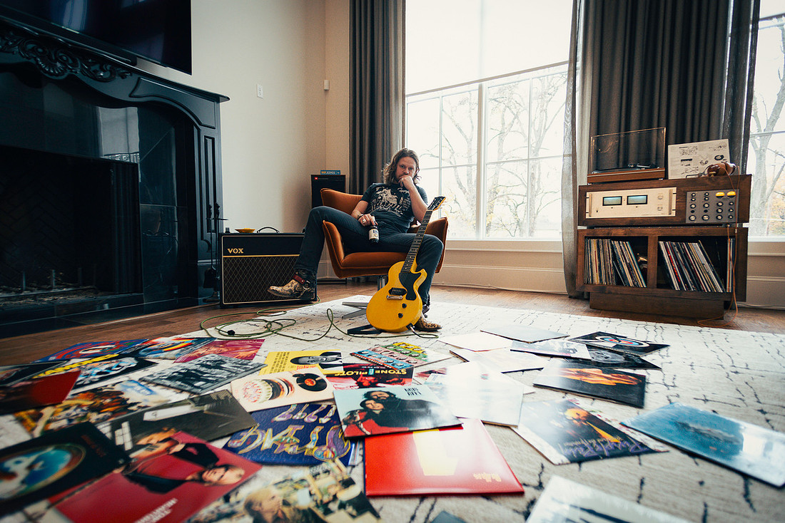  Sam Coburn sits in a mid-century modern chair with beer in one hand and guitar across his lap with his record collection sprawled all over the floor.